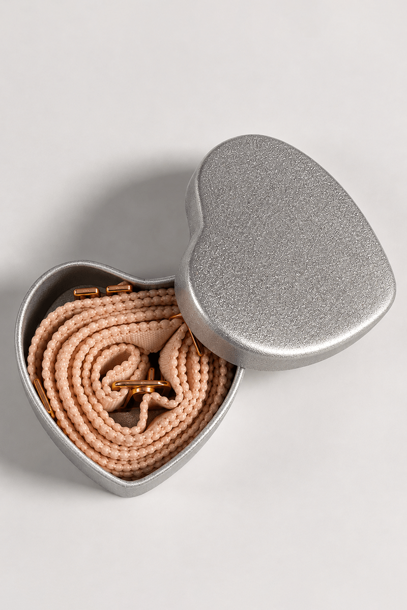 Close-up view of a pair of nude coloured ENDER LEGARD adjustable cross-over bra straps rolled in a heart-shaped silver presentation tin.