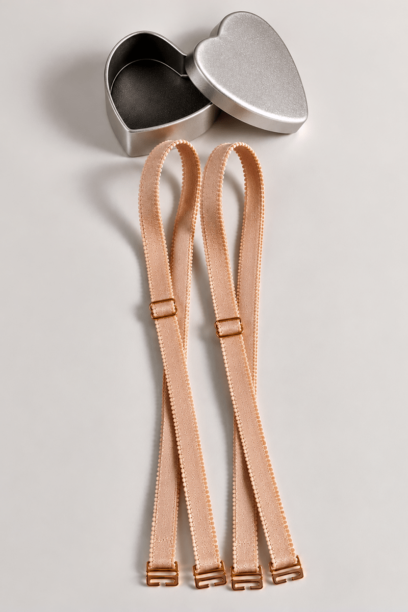 Close-up view of a pair of nude coloured ENDER LEGARD adjustable cross-over long bra straps next to a heart-shaped silver presentation tin.
