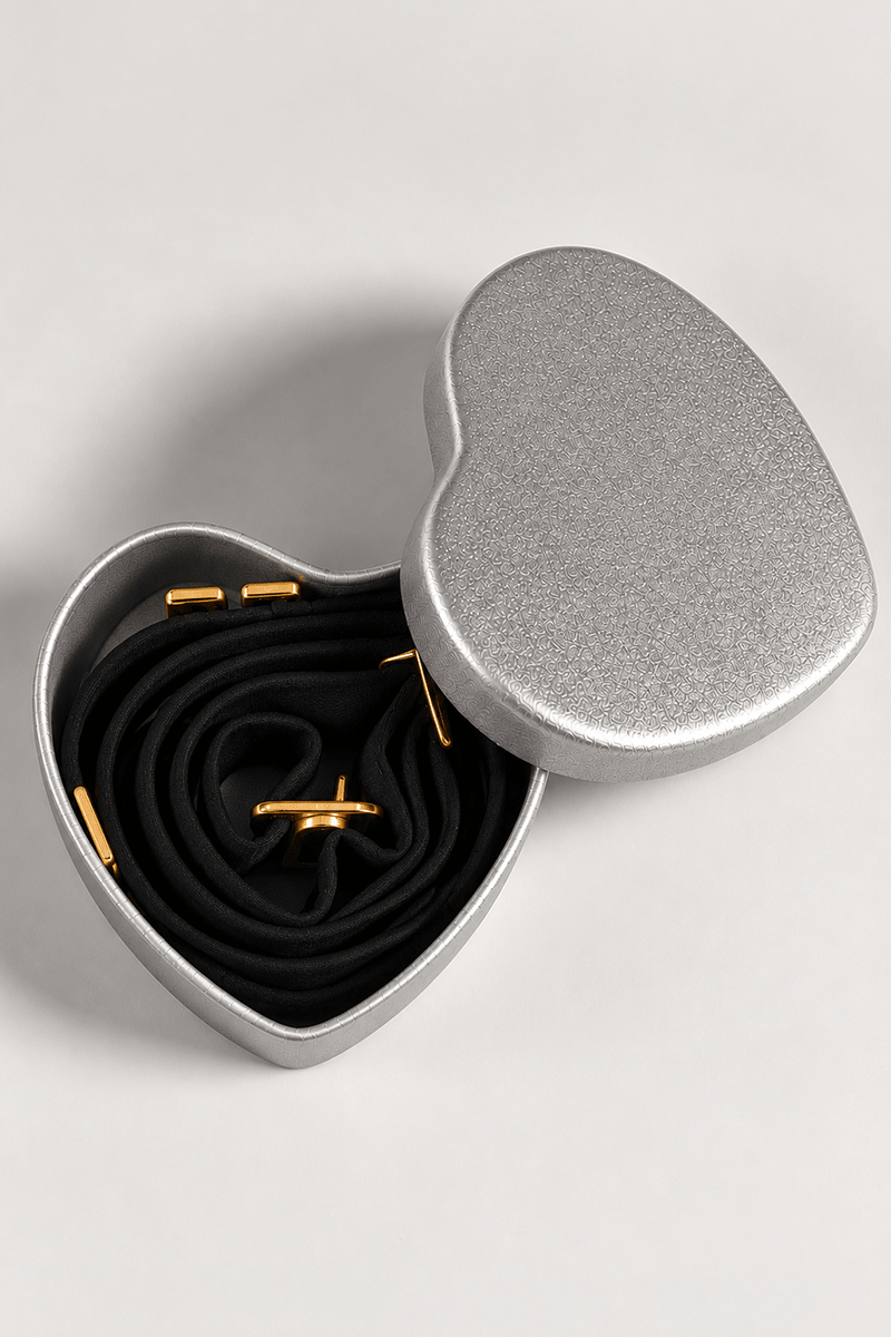 Close-up view of black adjustable cross-over bra straps with gold-toned fittings in a heart-shaped silver presentation tin.