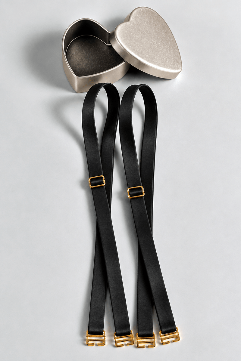 Close-up view of black cross-over long bra straps with gold-toned fittings and a heart-shaped silver presentation tin.