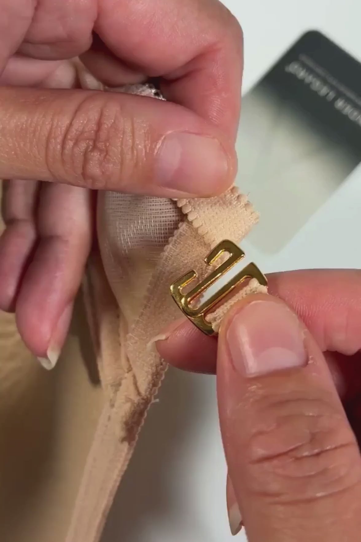 Close up on hands demonstrating how to fasten an Ender Legard Nude cross-over strap to a gold-toned G-hook to secure the strap to the bodysuit.